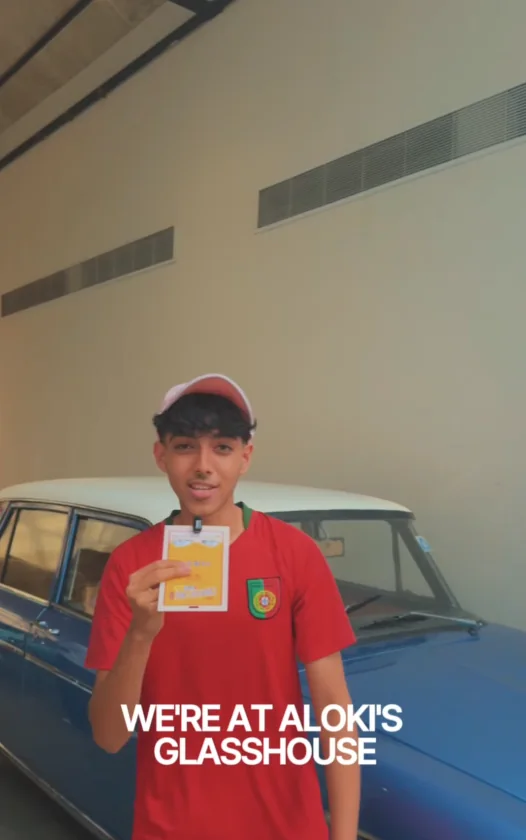 Presenter in a red Portugal shirt holding a ticket in front of a vintage blue car at Aloki's Glasshouse